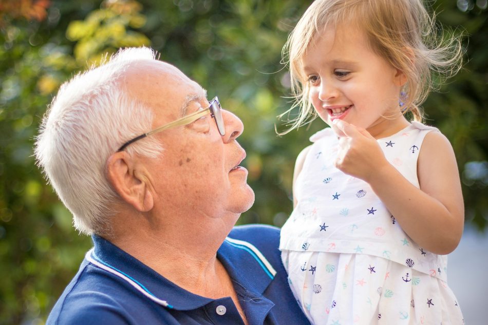 A grandfather holding his granddaughter in a garden, the two of them laughing.